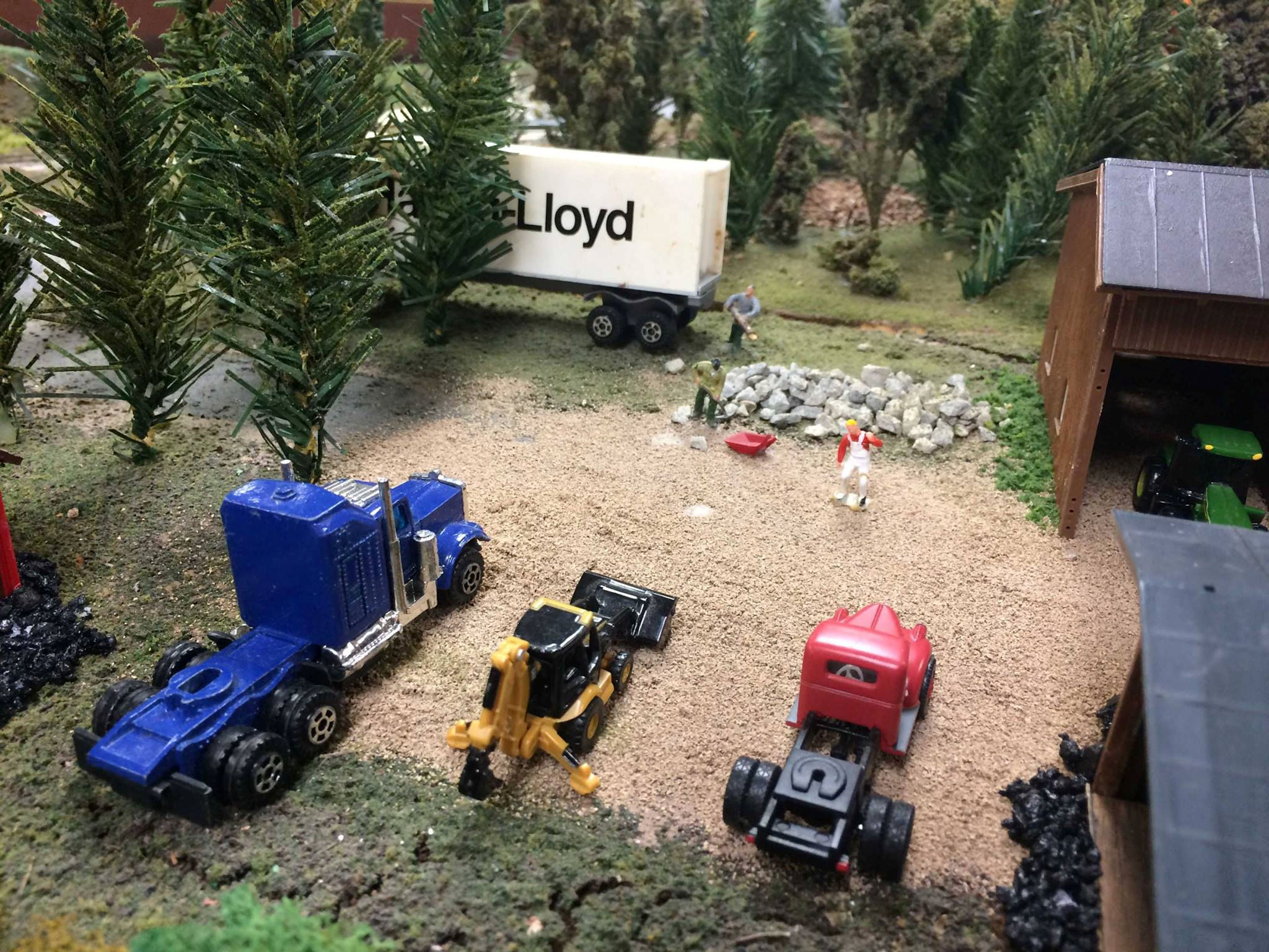 Model train layout featuring a truck loading area with miniature vehicles, trees, and workers, crafted by the Oshawa Pine Ridge Model Railroaders Club.