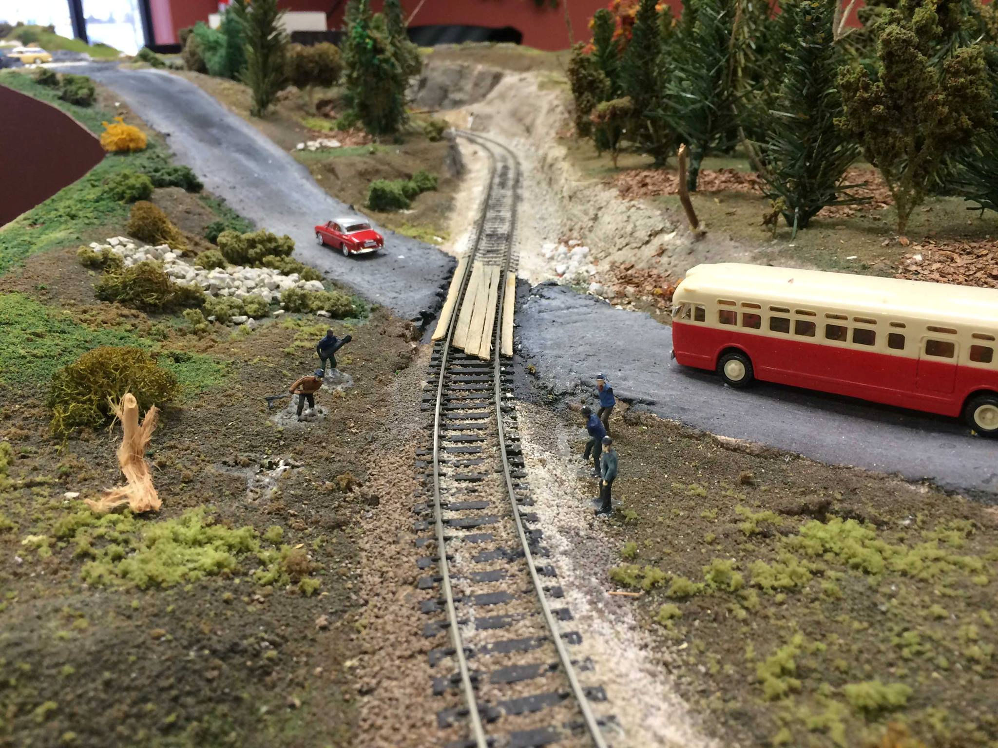 Miniature railway crossing with a red and cream vintage bus, train tracks, and figurines in a detailed model railroad landscape