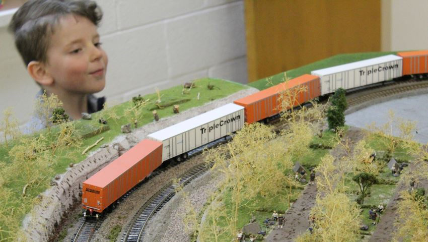 A child admires a model railroad scene featuring a Triple Crown RoadRailer train traveling through a miniature landscape with trees and hills.