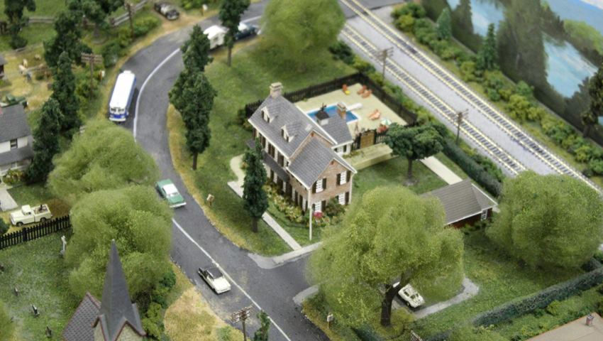 Miniature model railroad scene showcasing a suburban house, winding roads, trees, and a railway track.