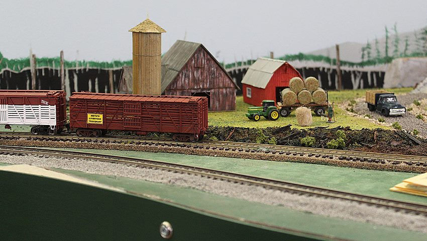 Model railroad farm scene with livestock train cars, red barns, a silo, hay bales, and farming equipment.