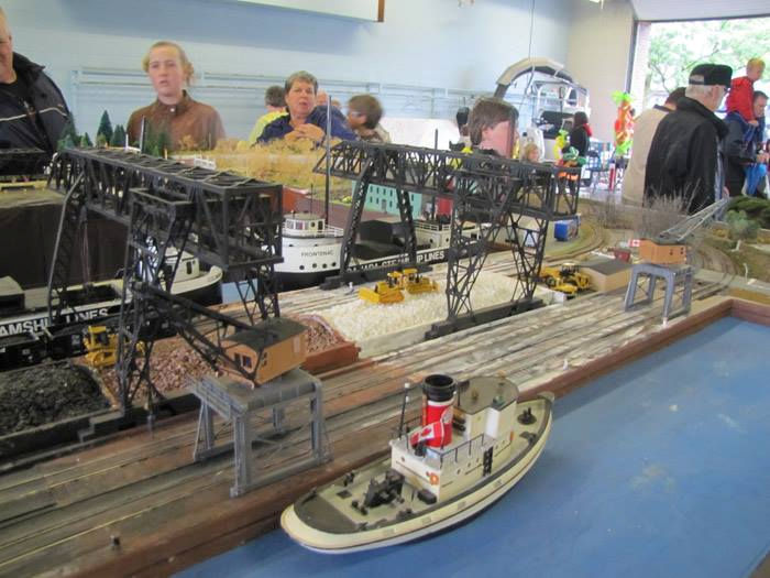 Model railroad display featuring a detailed waterfront scene with cargo ships, railcars, loading cranes, and a tugboat at a train club event.