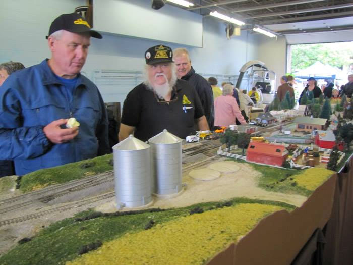 Model train enthusiasts observe a detailed rural train layout featuring grain silos, farm buildings, and scenic landscapes at a model railroading event.