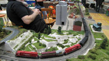 A freight train moving through an industrial model railway scene with warehouses and cranes.