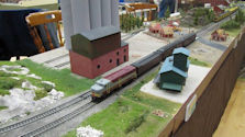 A passenger train at a detailed model railway station, featuring platform details and figures.