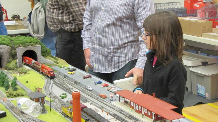 Young girl observing a model train set with a red station, a tunnel, and various locomotives at a model railroad exhibition.