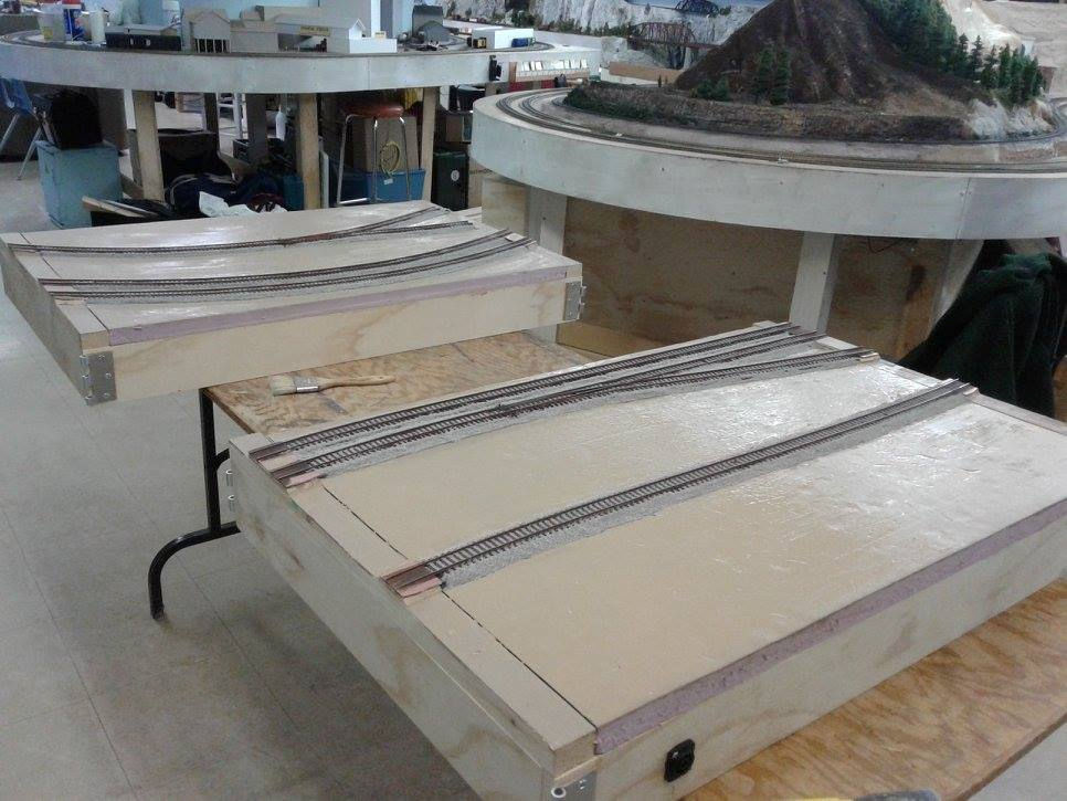 Two modular model railroad sections with freshly installed tracks, set on a worktable in a model train workshop.