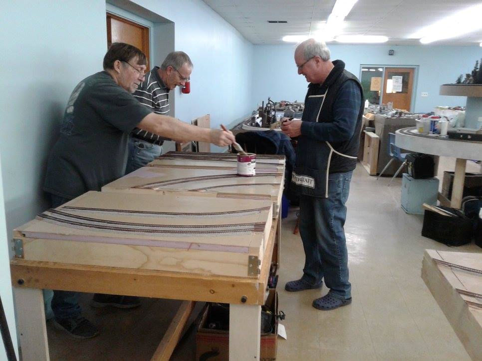 Three men working on a model railroad layout, painting and assembling track sections on a wooden frame.