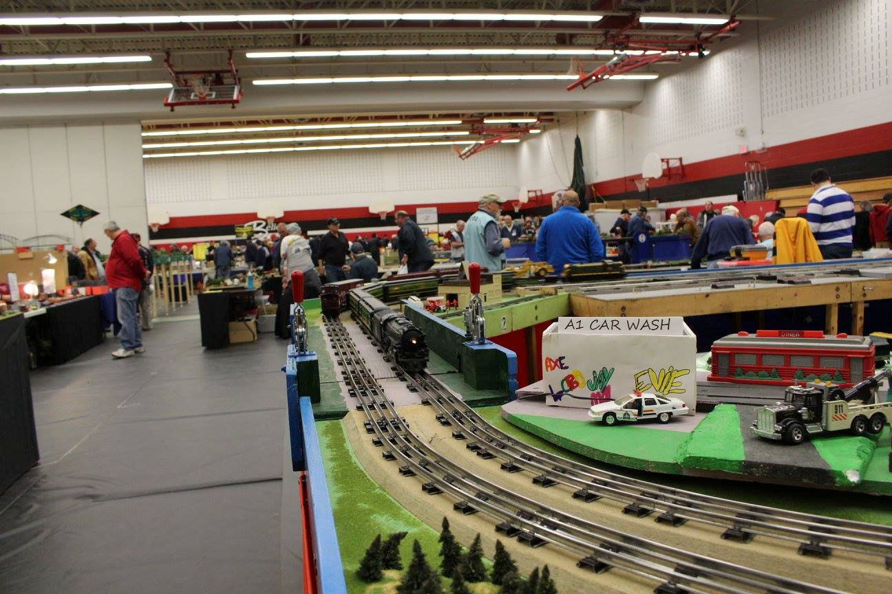 A model train layout with tracks, a steam locomotive, a diner, and a small handmade 'A1 Car Wash' sign, surrounded by visitors at a train exhibition.