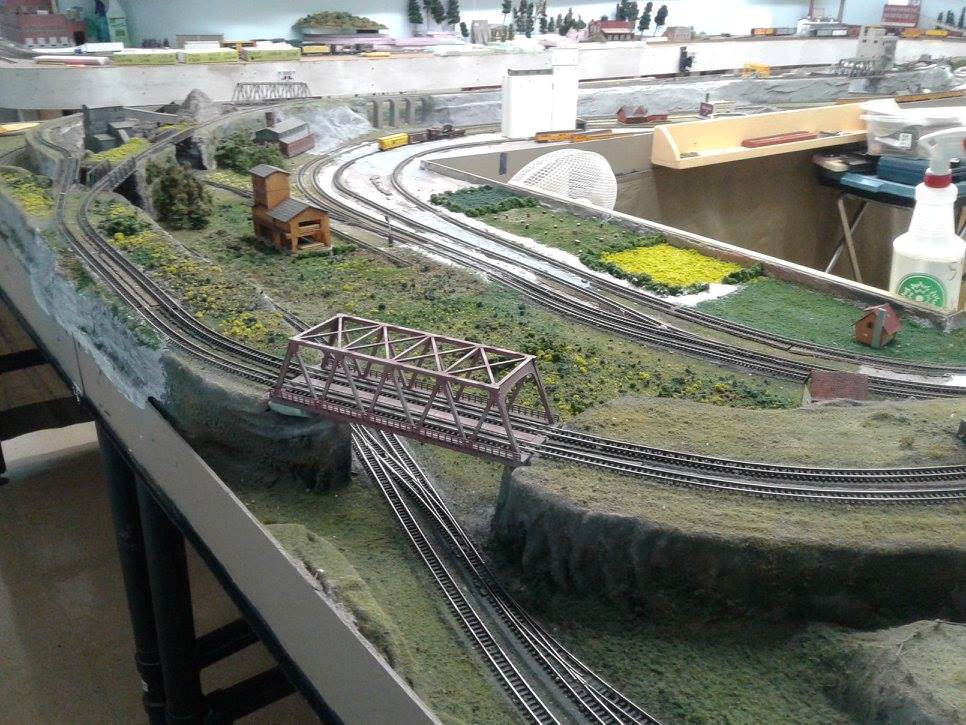 Model railroad layout with intersecting train tracks, a red truss bridge, and realistic scenery including grass, trees, and buildings.