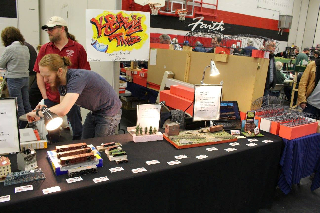 A model train vendor displaying custom weathered train cars, graffiti art, and scale structures at a train show.