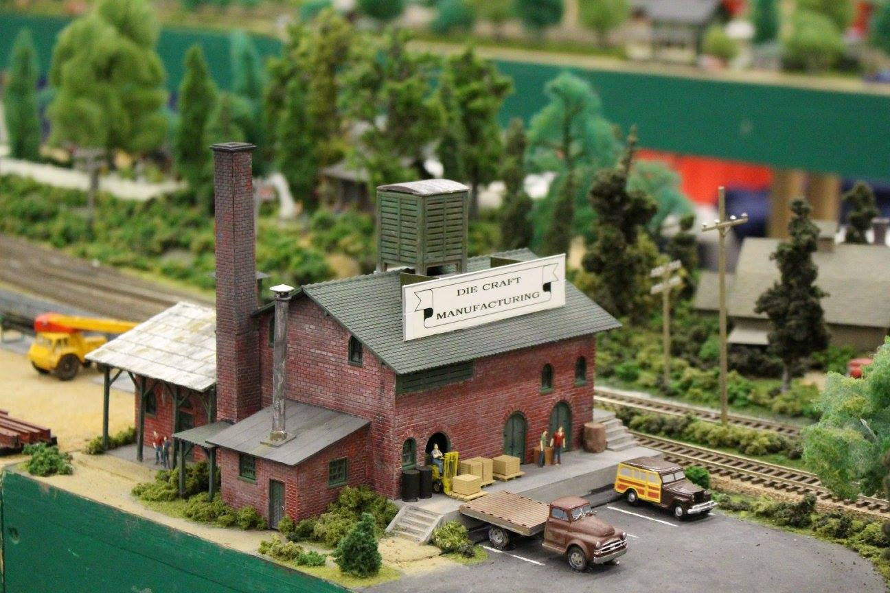 Miniature model of Die Craft Manufacturing, featuring a brick factory, parked trucks, workers, and railway tracks in a detailed model railroad layout.