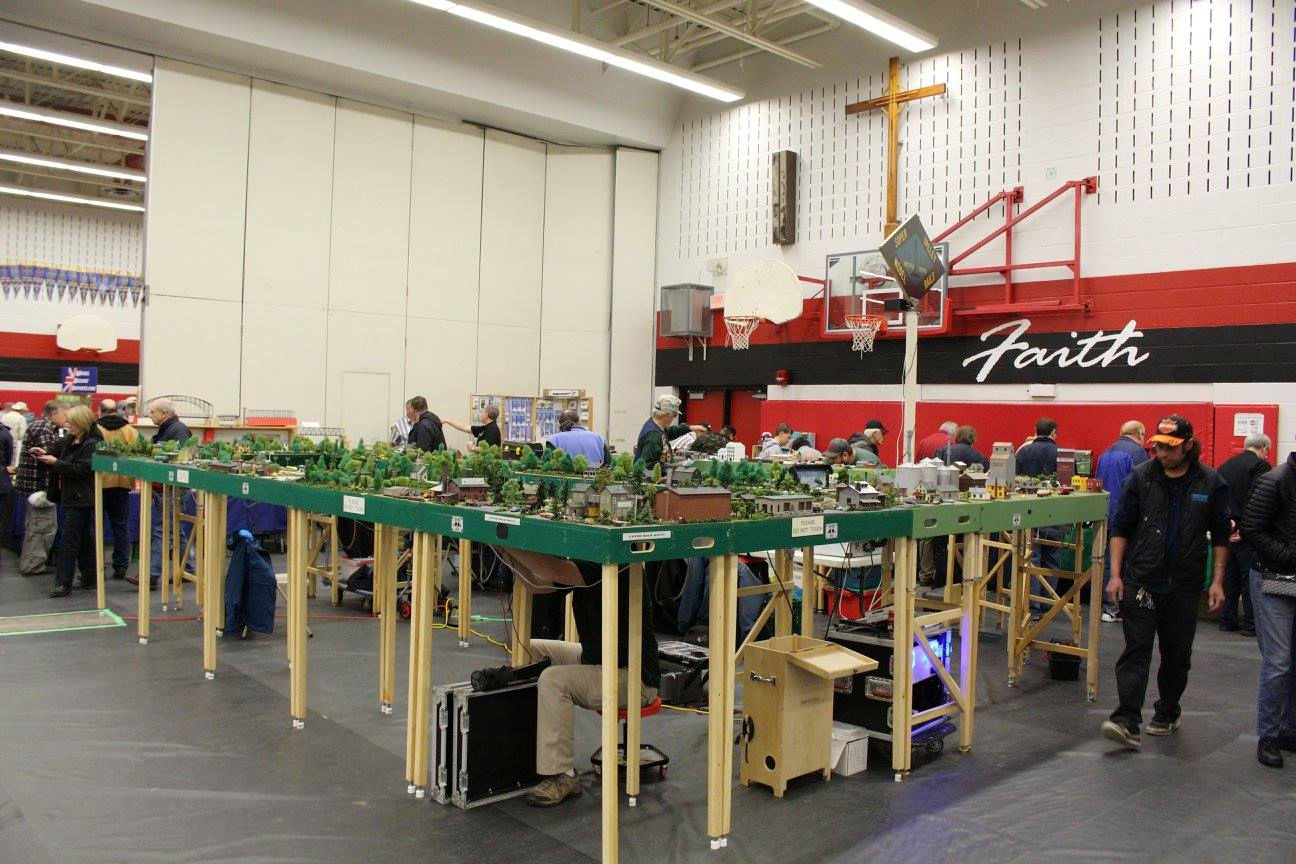 Large model railroad layout with detailed scenery, trees, and buildings at a train show, surrounded by hobbyists and collectors.