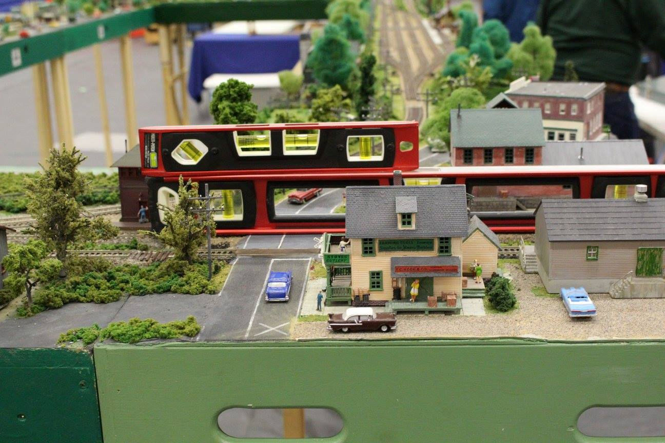 Miniature model town with buildings, classic cars, and a road crossing, with a red spirit level in the background.