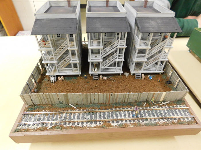 A model featuring three gray multi-story buildings with wooden staircases, a fenced yard, and a railway track in the foreground.